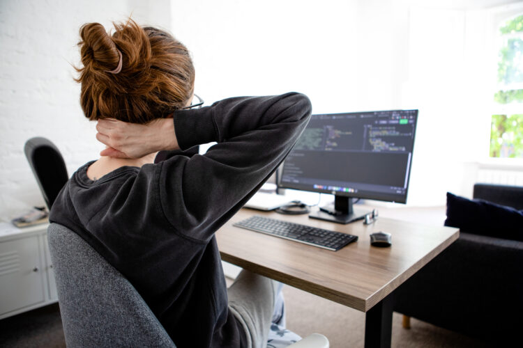 Una teletrabajadora con sudadera negra, gafas y moño se lleva la mano derecha al cuello mientras mira la pantalla de un ordenador.