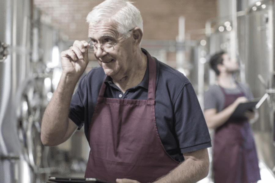 Hombre de unos 60 años con pelo blanco, gafas, camisa negra y delantal granate